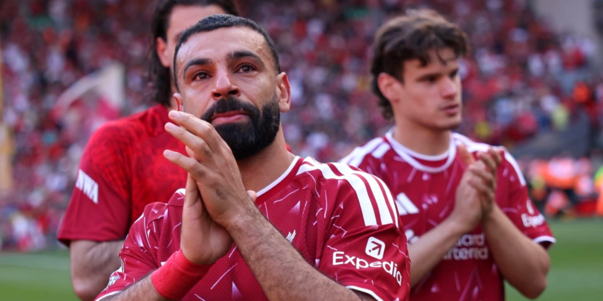 لقطة قريبة توثق وداع محمد صلاح لليفربول، حيث يرتدي القميص الأحمر، يبدو متأثراً بشدة وعيناه تدمعان، وهو يصفق بيديه مودعاً الجماهير في ملعب أنفيلد. يظهر يمين الإطار خلفه لاعب آخر من الفريق، مع خلفية لمدرجات مكتظة بالمشجعين