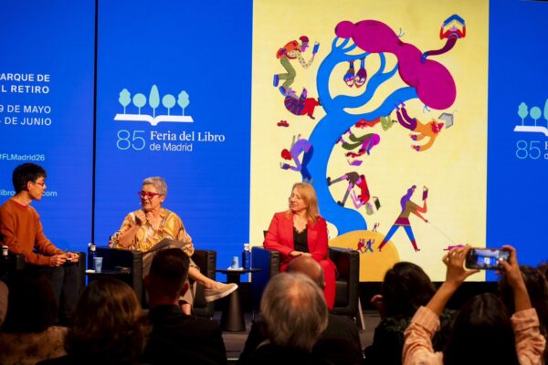 لقطة واسعة لمنصة المؤتمر التقديمي لـ معرض مدريد للكتاب 2026، حيث يجلس ثلاثة متحدثين (رجل وسيدتان) على كراسي سوداء، وتتحدث السيدة في المنتصف عبر الميكروفون. خلفهم شاشات عرض ضخمة زرقاء تحمل شعار الدورة 85، وتواريخ الانعقاد، ومكان الحدث في "ريتيرو"، وتتوسطها اللوحة الدعائية الرسمية ذات الخلفية الصفراء. في مقدمة الصورة، تظهر رؤوس وأيدي بعض الحضور وهم يلتقطون صوراً للمنصة بهواتفهم الذكية