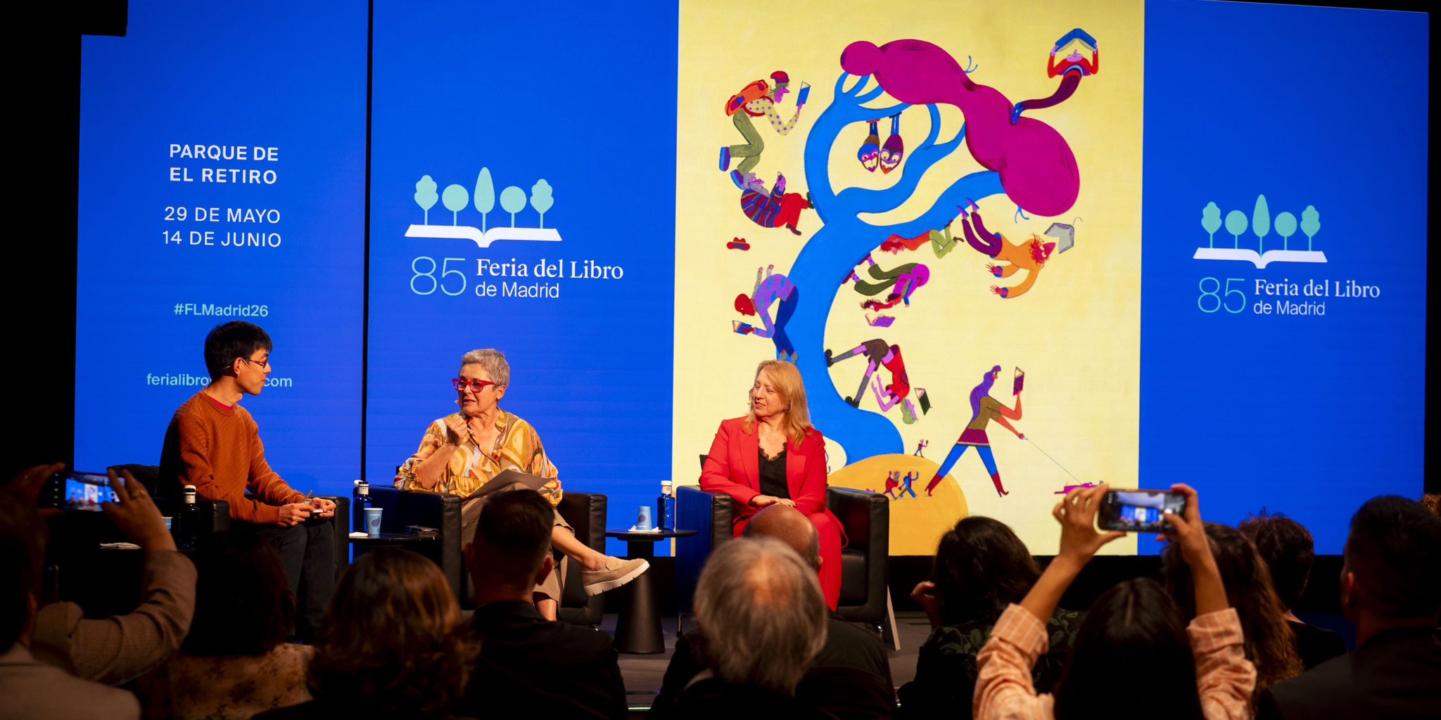 لقطة واسعة لمنصة المؤتمر التقديمي لـ معرض مدريد للكتاب 2026، حيث يجلس ثلاثة متحدثين (رجل وسيدتان) على كراسي سوداء، وتتحدث السيدة في المنتصف عبر الميكروفون. خلفهم شاشات عرض ضخمة زرقاء تحمل شعار الدورة 85، وتواريخ الانعقاد، ومكان الحدث في "ريتيرو"، وتتوسطها اللوحة الدعائية الرسمية ذات الخلفية الصفراء. في مقدمة الصورة، تظهر رؤوس وأيدي بعض الحضور وهم يلتقطون صوراً للمنصة بهواتفهم الذكية