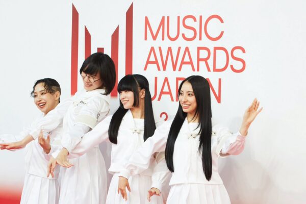 أربع فنانات يتألقن بأزياء بيضاء موحدة أثناء وقوفهن أمام شعار فعاليات جوائز MUSIC AWARDS JAPAN 2026