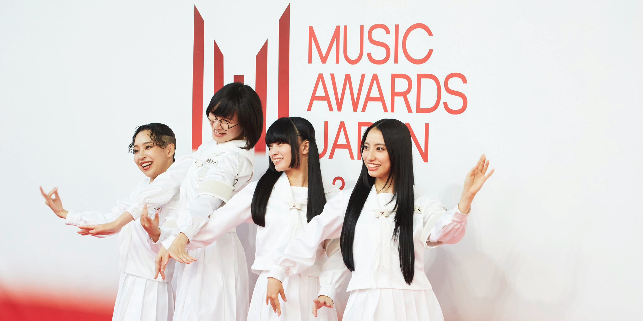 أربع فنانات يتألقن بأزياء بيضاء موحدة أثناء وقوفهن أمام شعار فعاليات جوائز MUSIC AWARDS JAPAN 2026