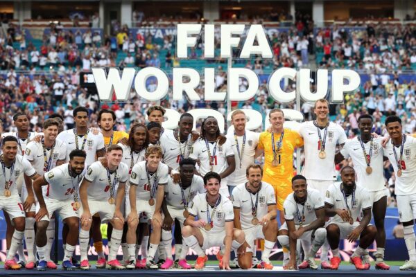 صورة جماعية احتفالية لمنتخب إنجلترا لكرة القدم على منصة في ملعب مزدحم، بعد الفوز بالميداليات البرونزية في كأس العالم FIFA 2026. يرتدي اللاعبون والمدرب زي إنجلترا الأبيض وميداليات برونزية. علامة كبيرة ثلاثية الأبعاد تحمل نص "FIFA WORLD CUP 2026" مرئية في الخلفية