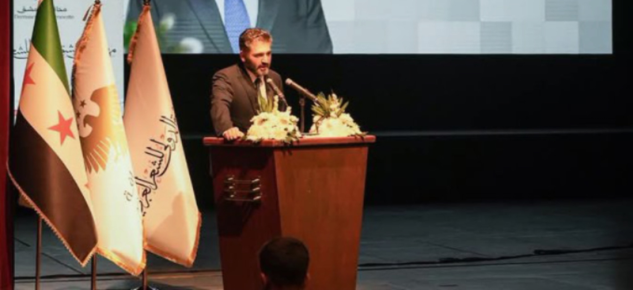وزير الثقافة محمد ياسين الصالح يلقي الكلمة الافتتاحية من على المنصة الرسمية لفعاليات مهرجان دمشق للشعر العربي، وتظهر بجانبه الأعلام الرسمية ومنها العلم السوري