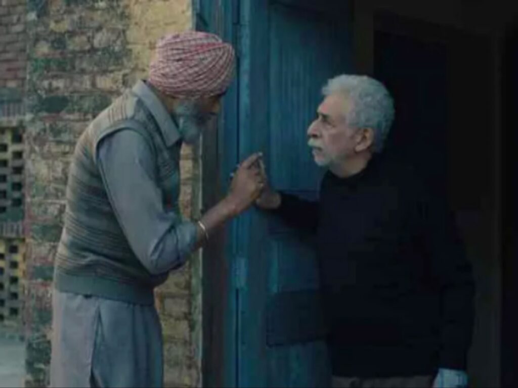 مشهد سينمائي يجمع بين النجم نصير الدين شاه ورجل سيخي مسن في حوار هادئ ونظرات متبادلة أمام باب خشبي أزرق، مما يعكس التفاعل الإنساني العميق وسط الانقسامات