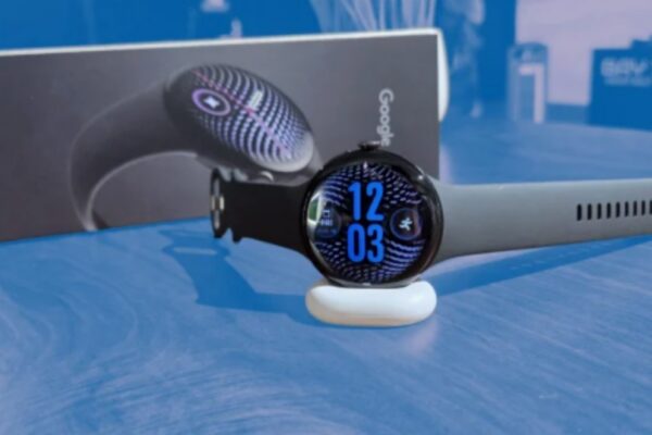 ساعة Pixel Watch 5 الذكية باللون الأسود مستقرة على قاعدة شحن بيضاء وخلفها عبوة التغليف الأصلية