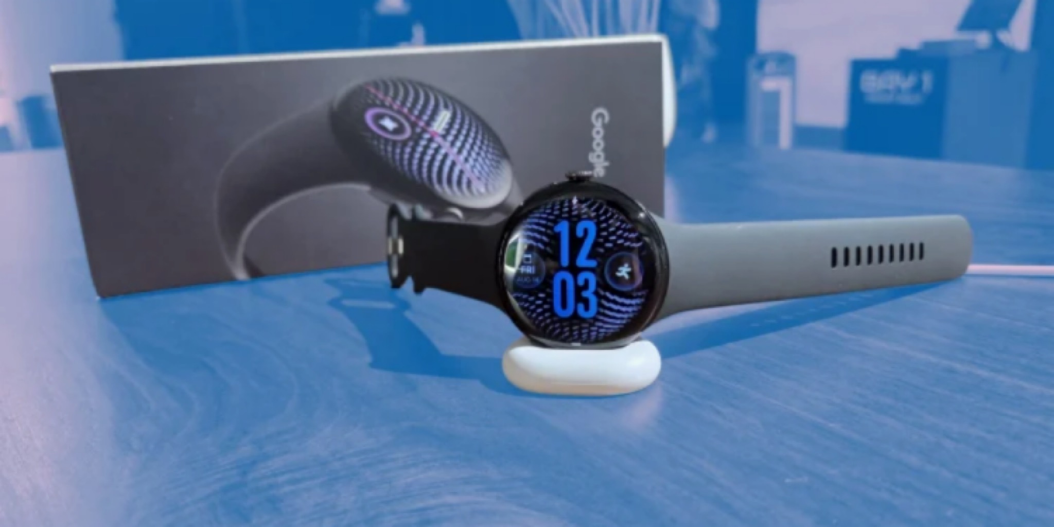 ساعة Pixel Watch 5 الذكية باللون الأسود مستقرة على قاعدة شحن بيضاء وخلفها عبوة التغليف الأصلية