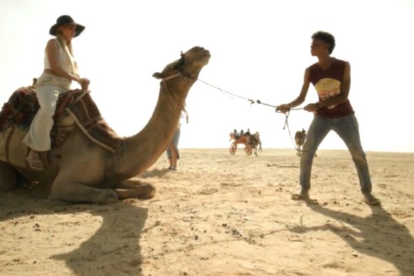 لقطة نهارية من فيلم خوفو Khufu تظهر الصبي سيد وهو يشد بقوة حبل جمل بارك على رمال الصحراء، بينما تجلس فوقه سائحة أجنبية