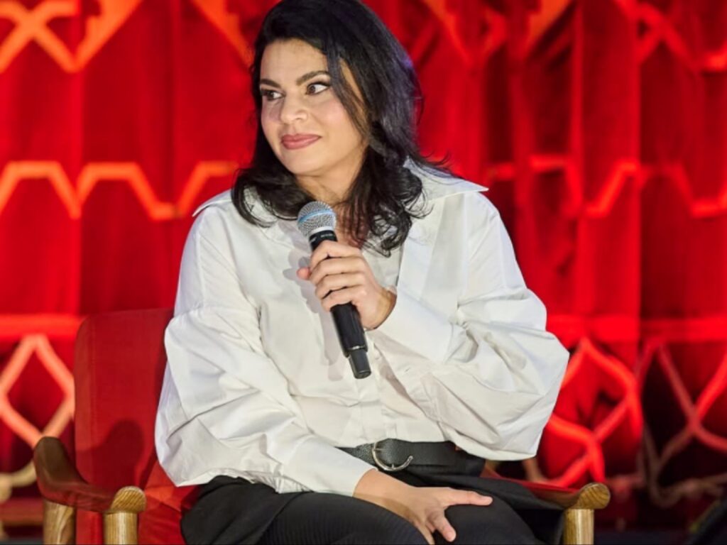 فنانة وكاتبة رومانية تتحدث عبر الميكروفون خلال ندوة فنية مرتدية قميصاً أبيض أنيقاً