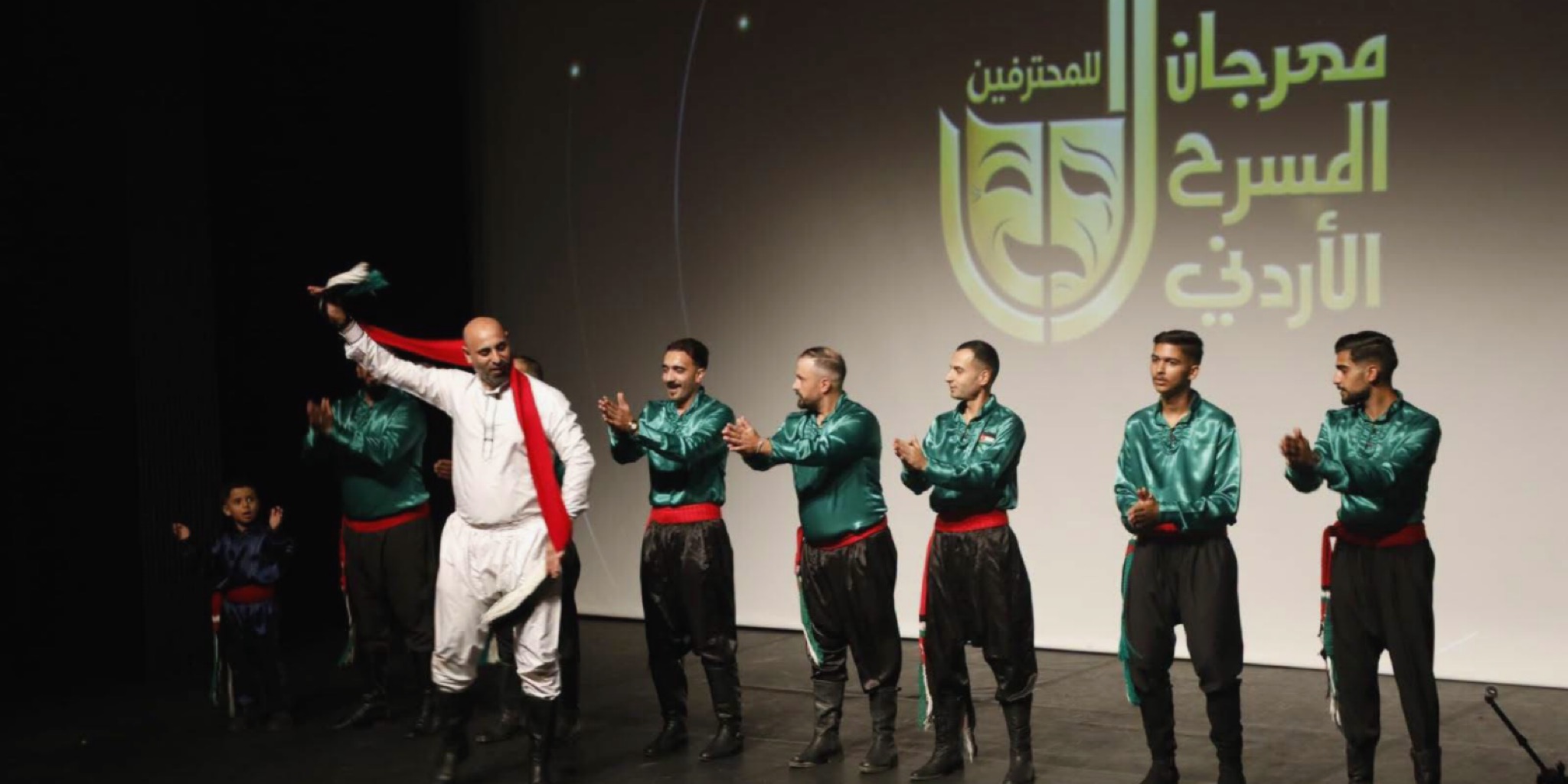 فرقة الرمثا للفنون الشعبية تؤدي عرضاً فلكلورياً مميزاً على خشبة المسرح، حيث يرتدي الأعضاء أزياء تقليدية خضراء وسوداء ويقفون في صف واحد يصفقون، بينما يظهر في الخلفية الشعار الذهبي الضخم الذي يحمل اسم مهرجان المسرح الأردني للمحترفين