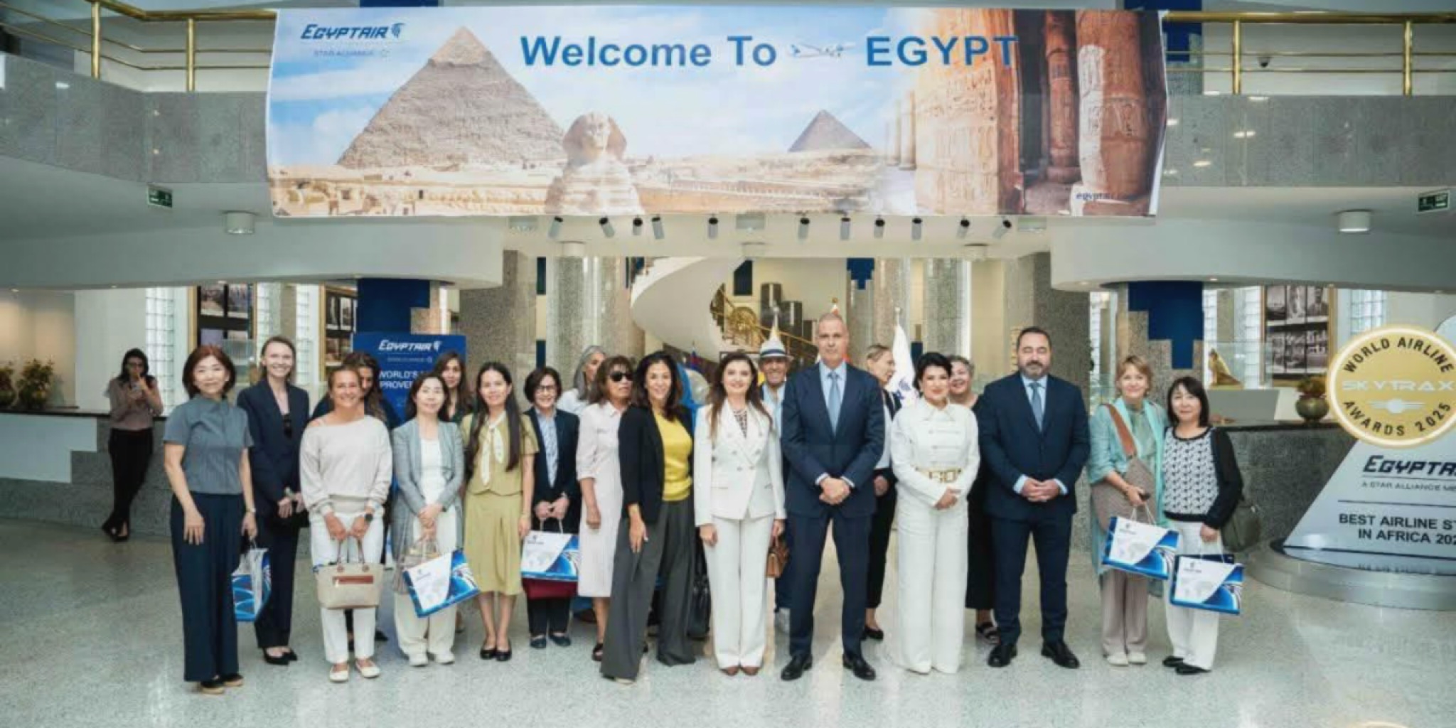 الطيار أحمد عادل يتوسط وفد زوجات الدبلوماسيين الأجانب في صورة تذكارية جماعية داخل المبنى الرئيسي التابع لقطاعات الشركة القابضة لمصر للطيران، ويظهر في الخلفية لافتة ترحيبية تحمل عبارة (Welcome To EGYPT) وصوراً للأهرامات والمعالم الأثرية، وبجوارهم درع يمثل جائزة سكاي تراكس العالمية
