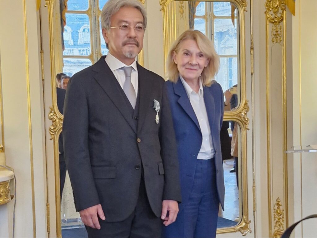صورة تذكارية تجمع إيجي أنوما بسيدة داخل قاعة مزينة بالمرايا والزخارف الذهبية الكلاسيكية في مقر وزارة الثقافة بباريس، ويظهر المبدع الياباني مرتدياً بزة رسمية داكنة تتزين بشارة وسام فارس الفنون والآداب على صدره