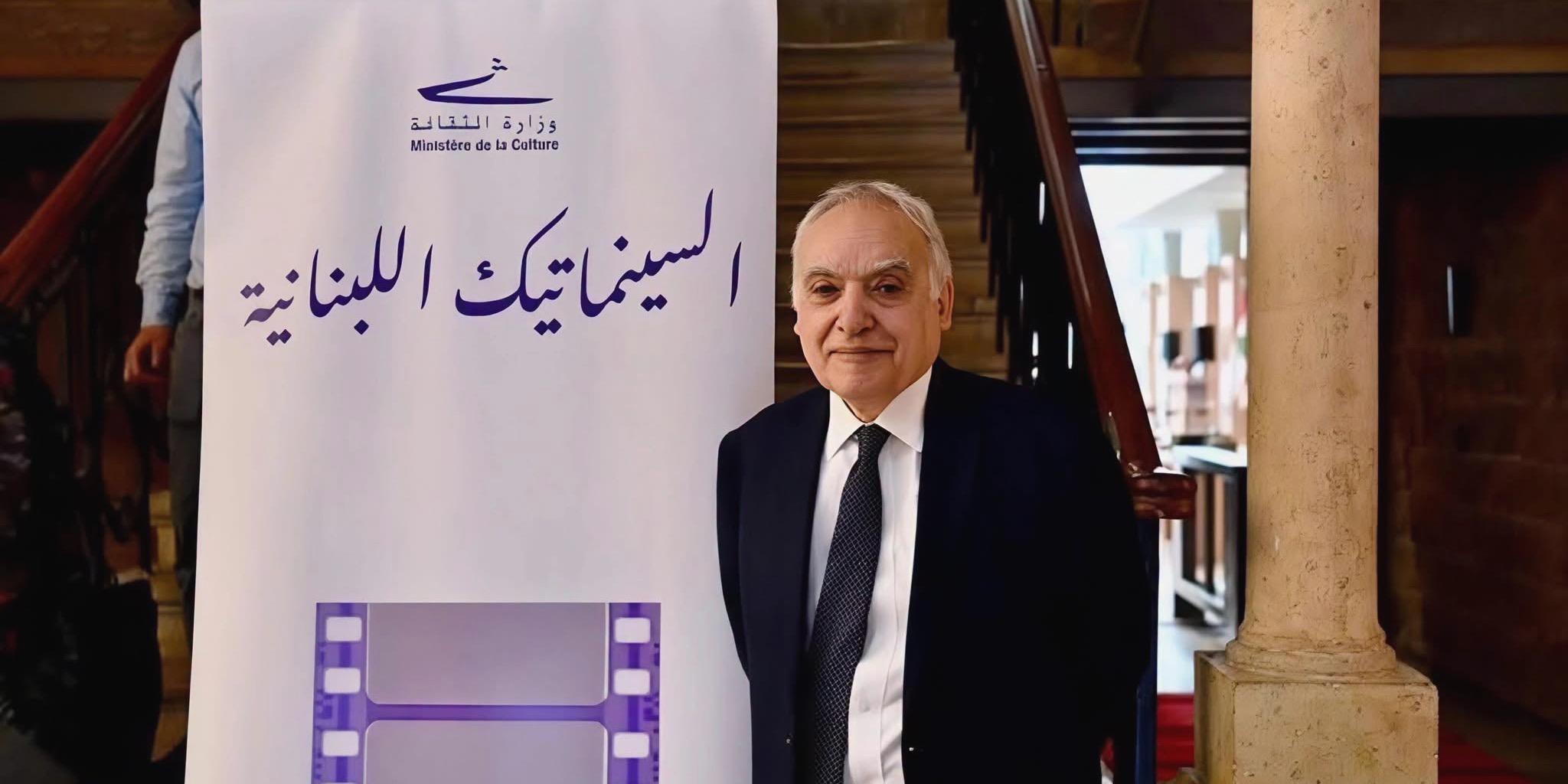 وزير الثقافة اللبناني غسان سلامة مرتدياً بدلة رسمية داكنة مع ربطة عنق، يقف بجانب لافتة بيضاء تحمل شعار الوزارة وعبارة "السينماتيك اللبنانية" في حفل افتتاح تظاهرة أسبوع السينما اللبنانية